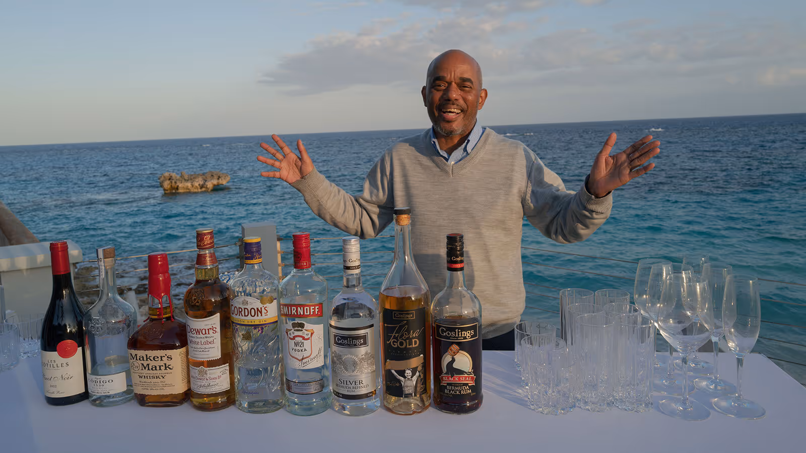 A man stands behind a table with various liquor bottles and empty glasses by the ocean.