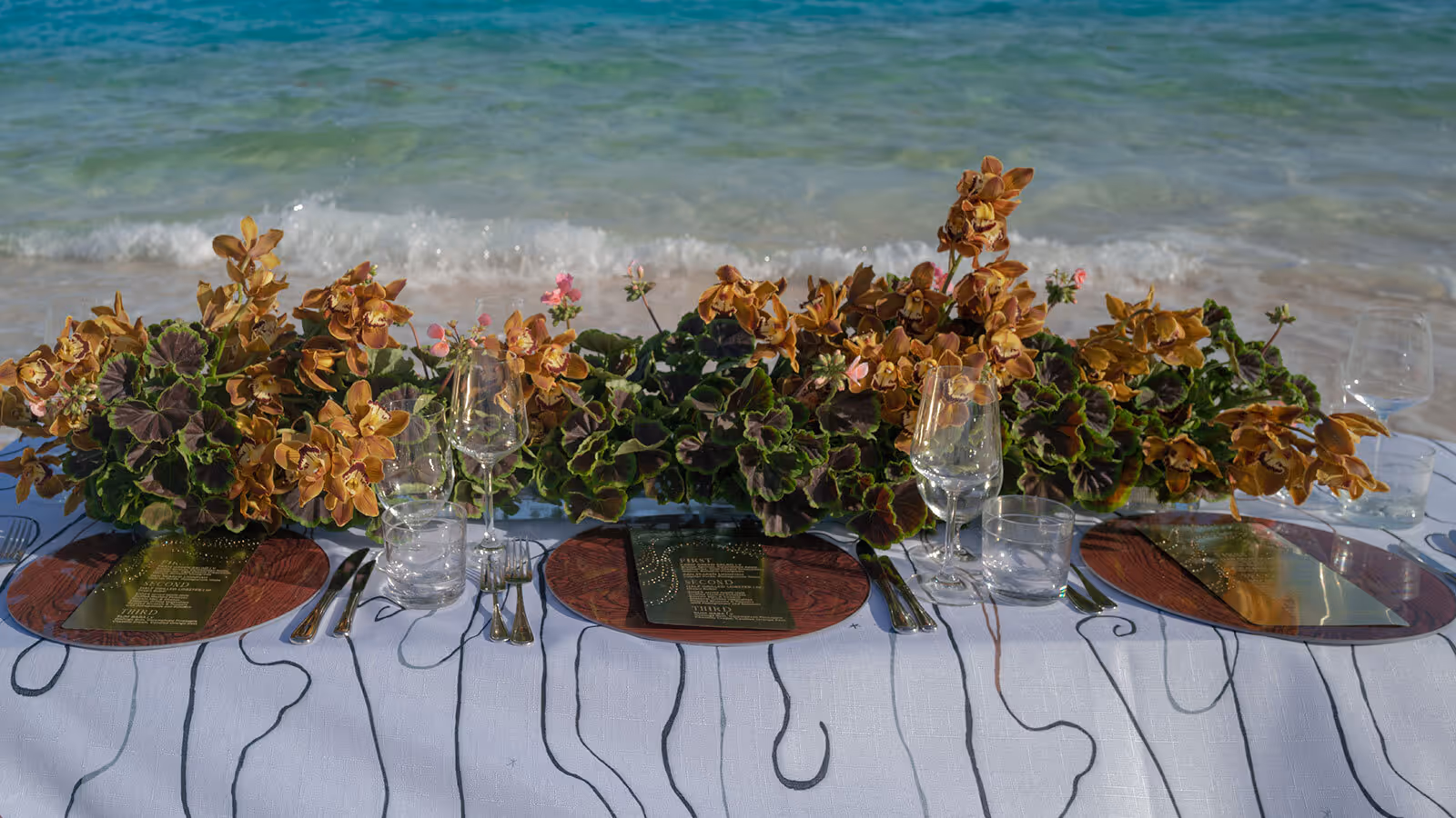 A table set with floral centerpieces and glassware is positioned by the shore with waves in the background.