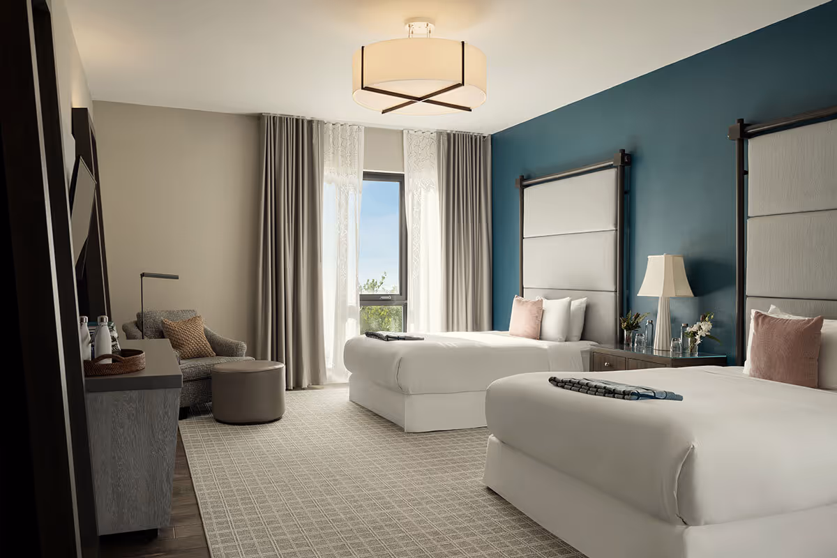 Modern hotel room with two twin beds, a blue accent wall, armchair, TV, and large window with curtain.