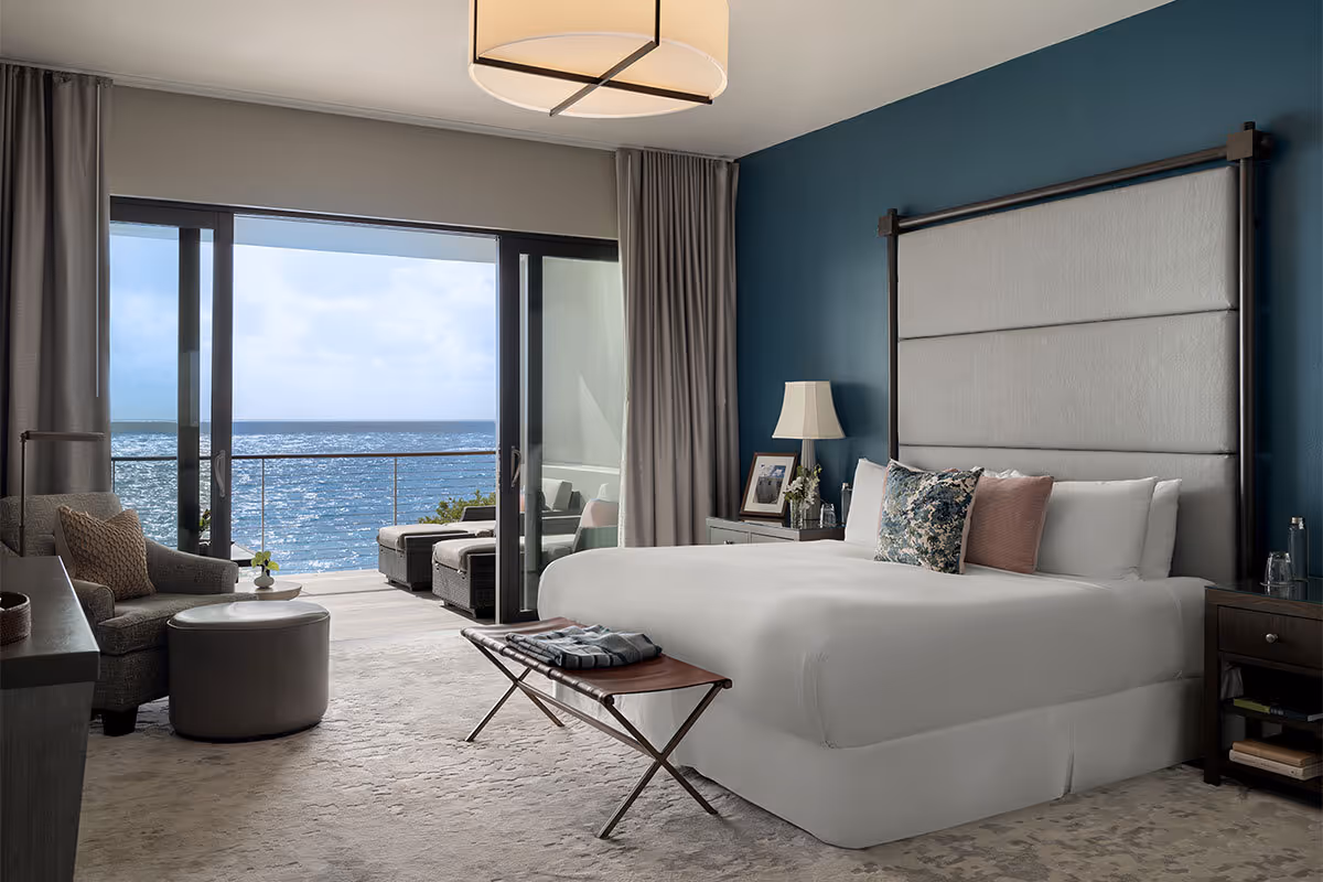 Modern hotel room with a large bed, seating area, and balcony overlooking the ocean through glass doors.