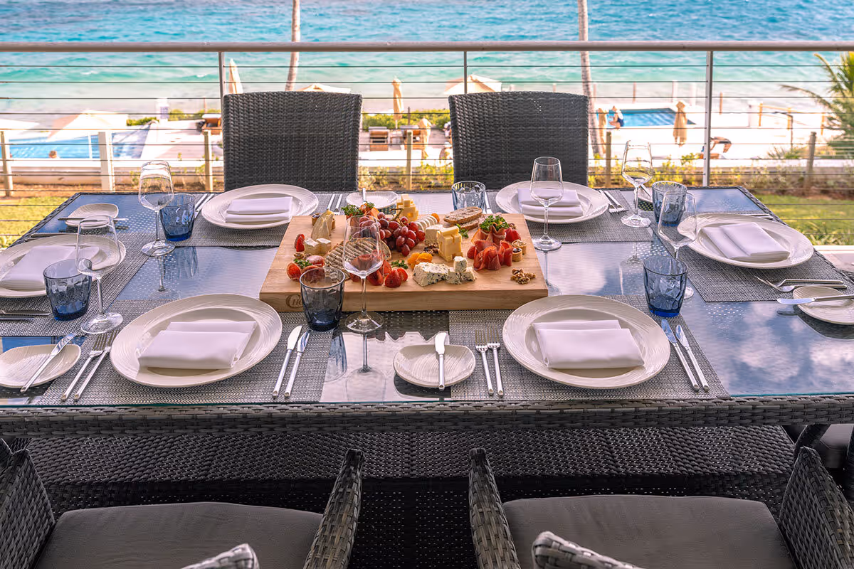 Outdoor dining table set for six with plates, cutlery, glasses, and a cheese platter, overlooking the sea.