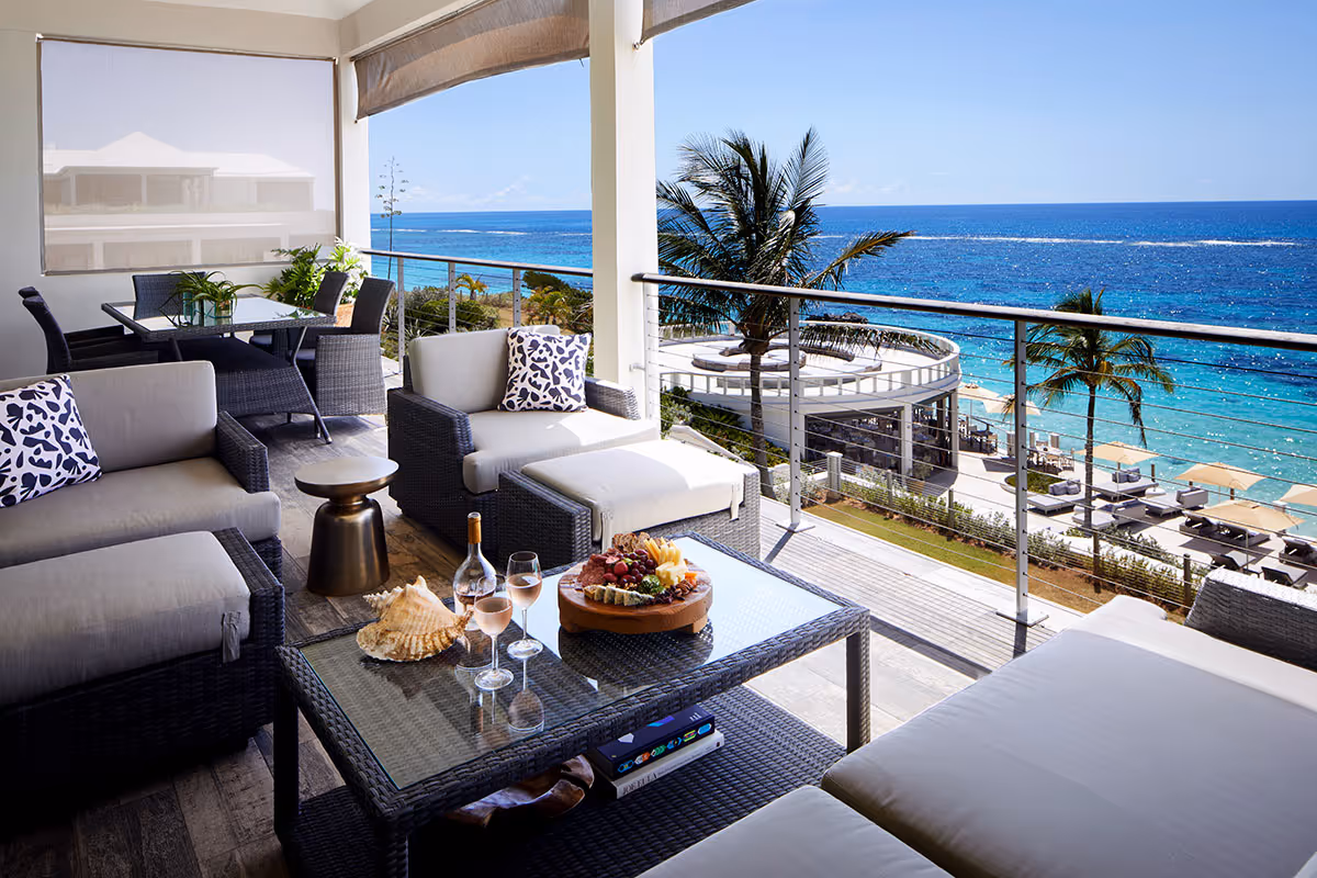 Modern balcony with lounge seating, snacks, and drinks, overlooking a pool area and the ocean on a sunny day.