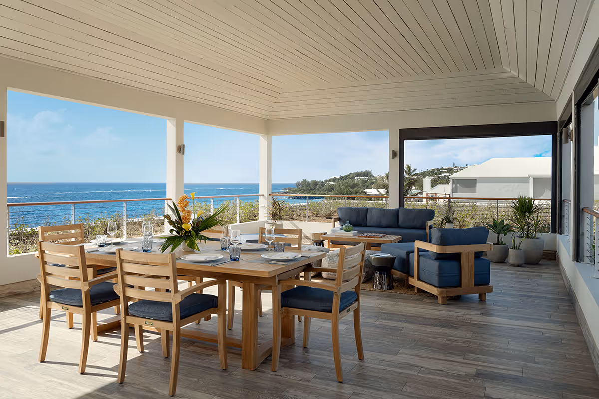 Covered patio with wooden dining and lounge furniture, set for a meal, overlooking the ocean and coastal landscape.
