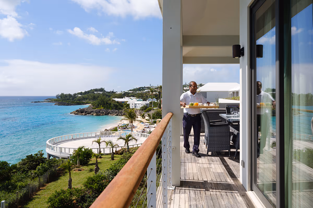 A man stands on a balcony by the ocean, holding a tray with drinks, surrounded by outdoor furniture and coastal scenery.