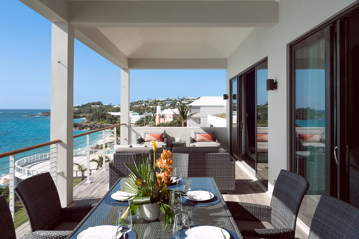 Covered balcony with dining table and lounge area overlooks the ocean and coastal houses under a clear blue sky.