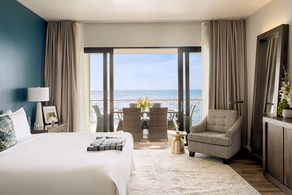 A hotel room with a bed, armchair, and balcony overlooking the ocean with a dining table and chairs outside.