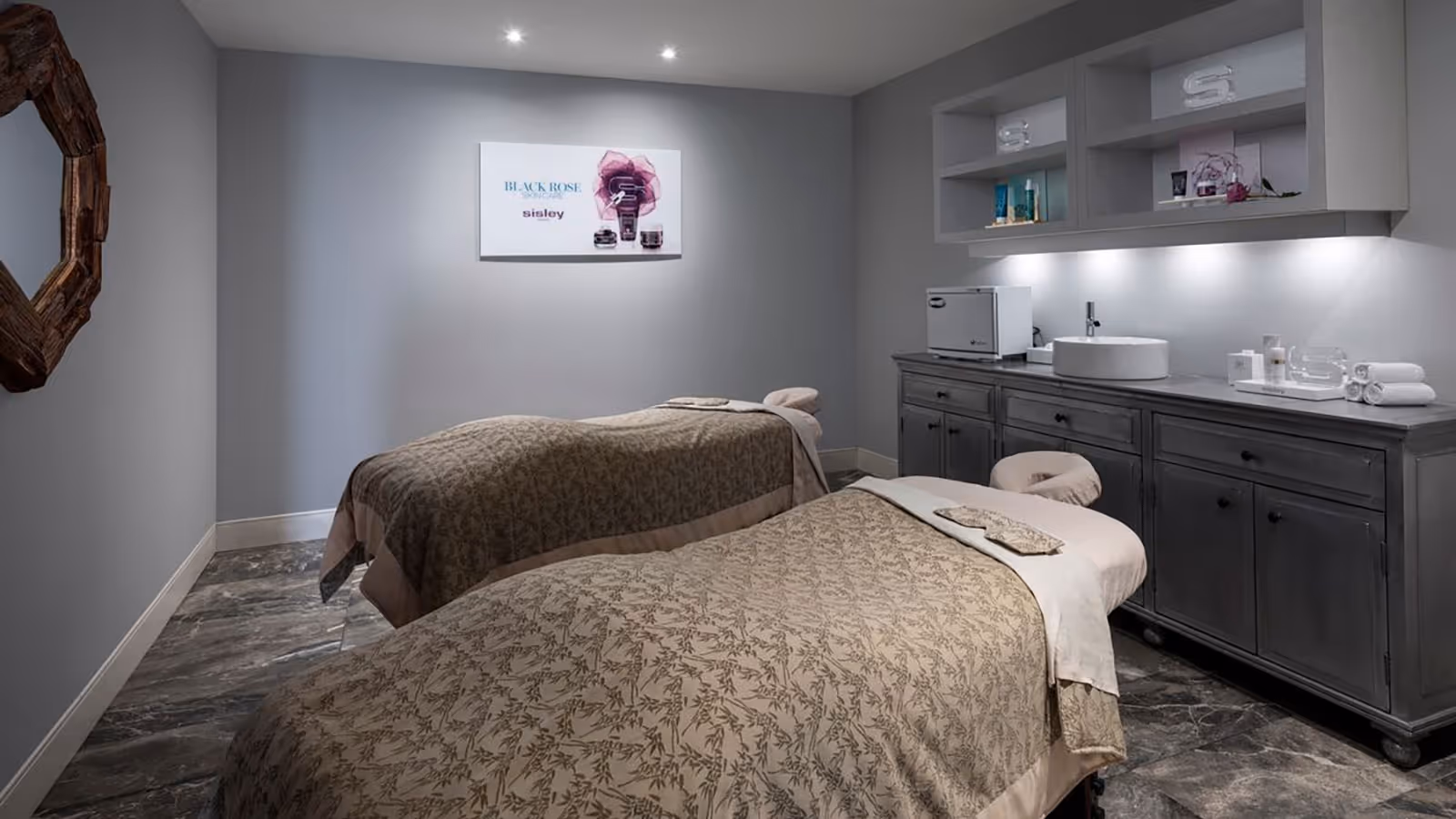Spa treatment room with two massage tables covered in patterned blankets and a counter with sink and skincare products.