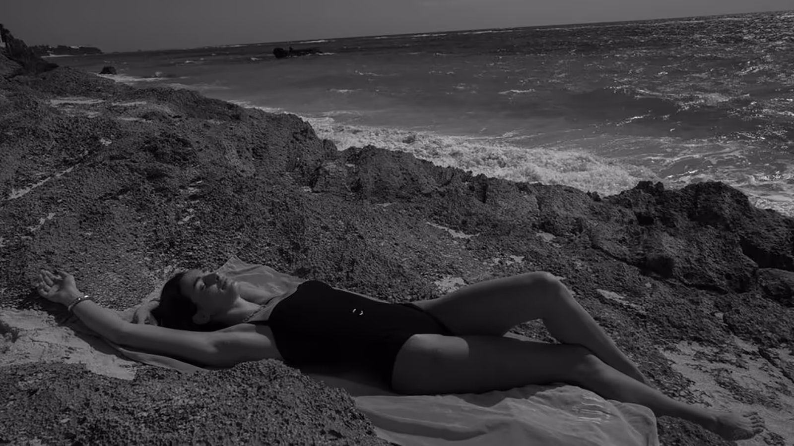 Woman in a black swimsuit lying on a towel on rocky shore with ocean waves in the background.