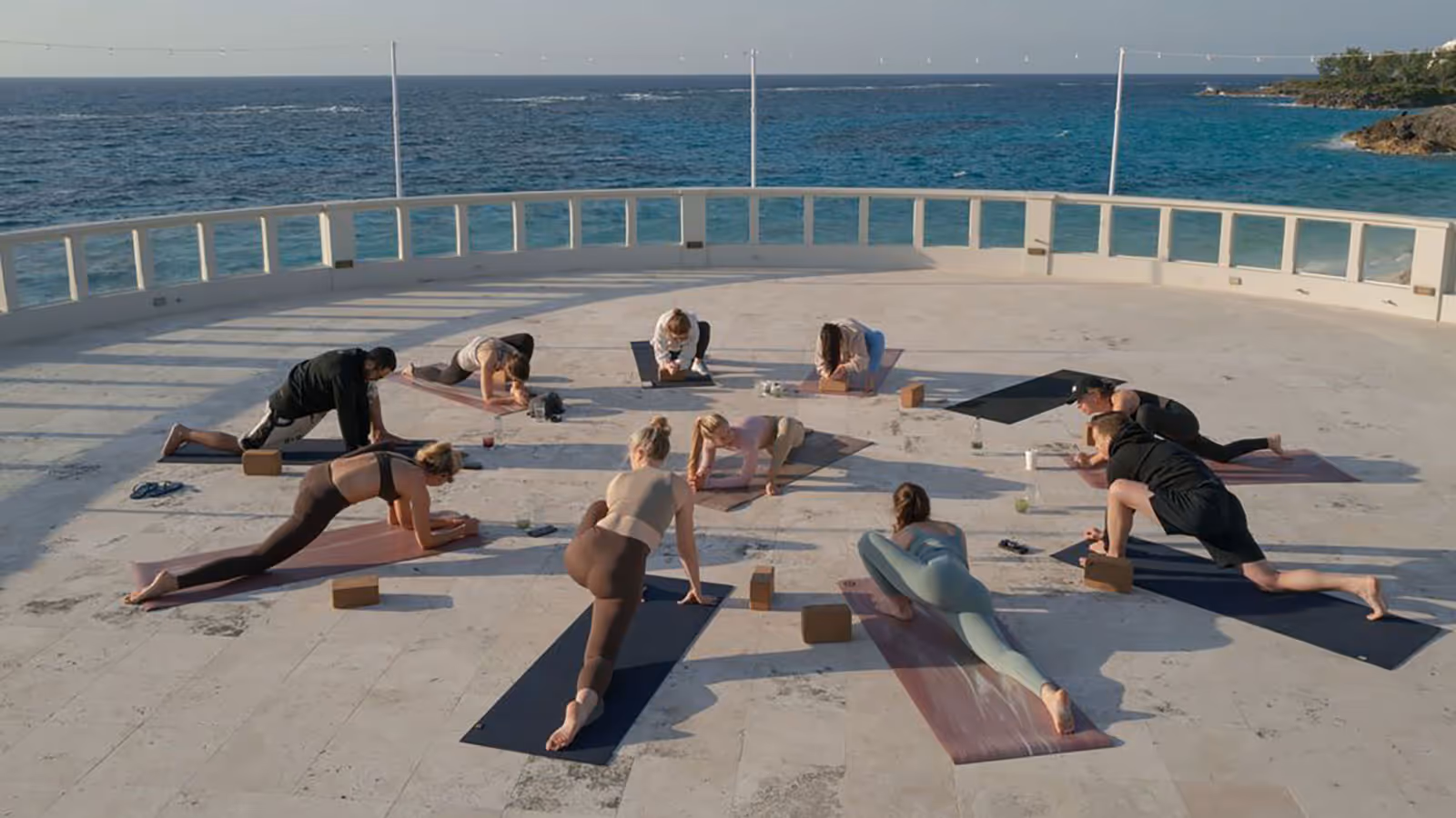 Group of people practicing yoga poses on mats outdoors on a terrace overlooking the ocean.