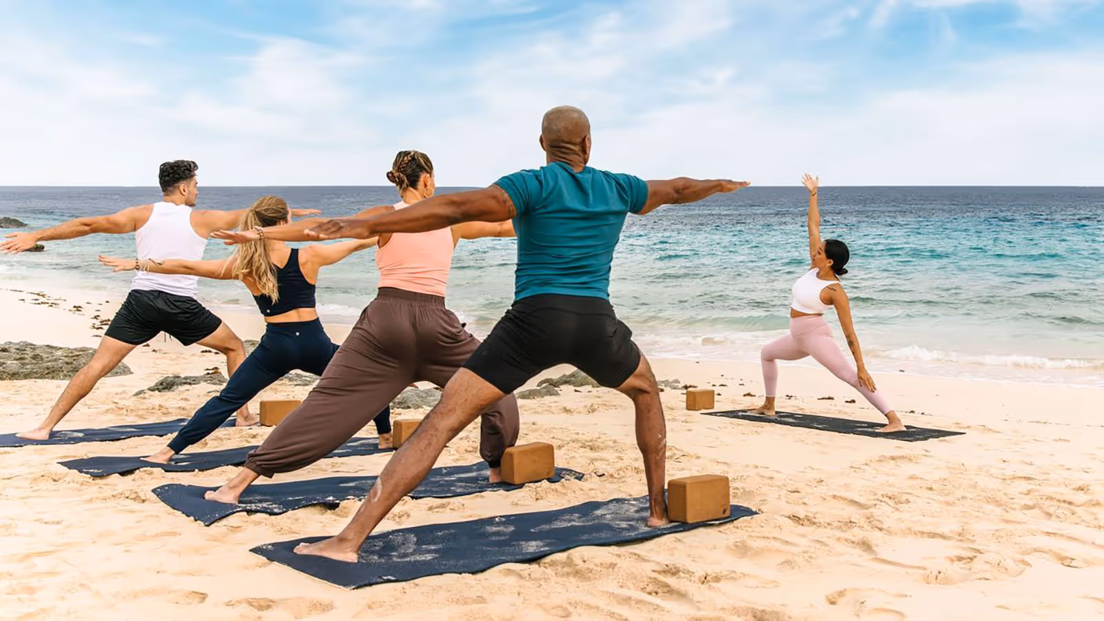 Group of five people practicing yoga on mats on a sandy beach near the ocean, performing warrior poses.