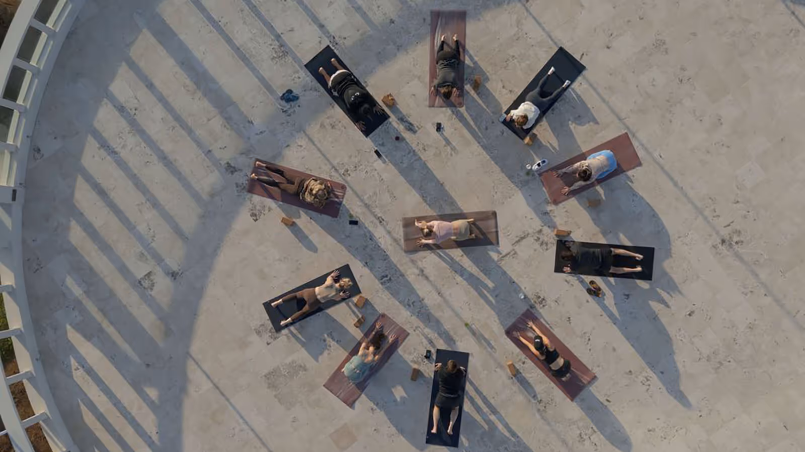 Aerial view of ten people lying on yoga mats arranged in a circle on a concrete surface, engaging in a group yoga session outdoors.