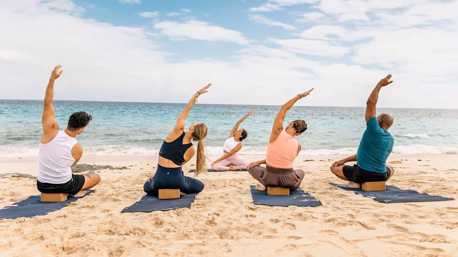 Group of five people doing seated side stretches on yoga mats on a sandy beach by the ocean.