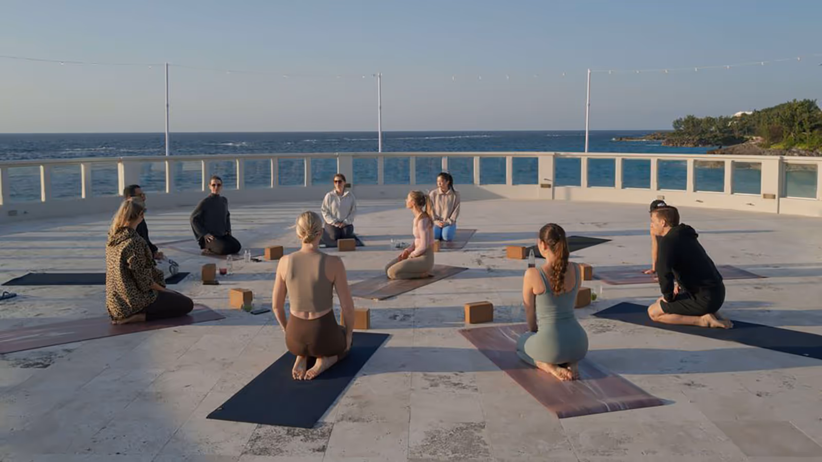 Group of people sitting on yoga mats in a circle outdoors by the ocean during a yoga session.