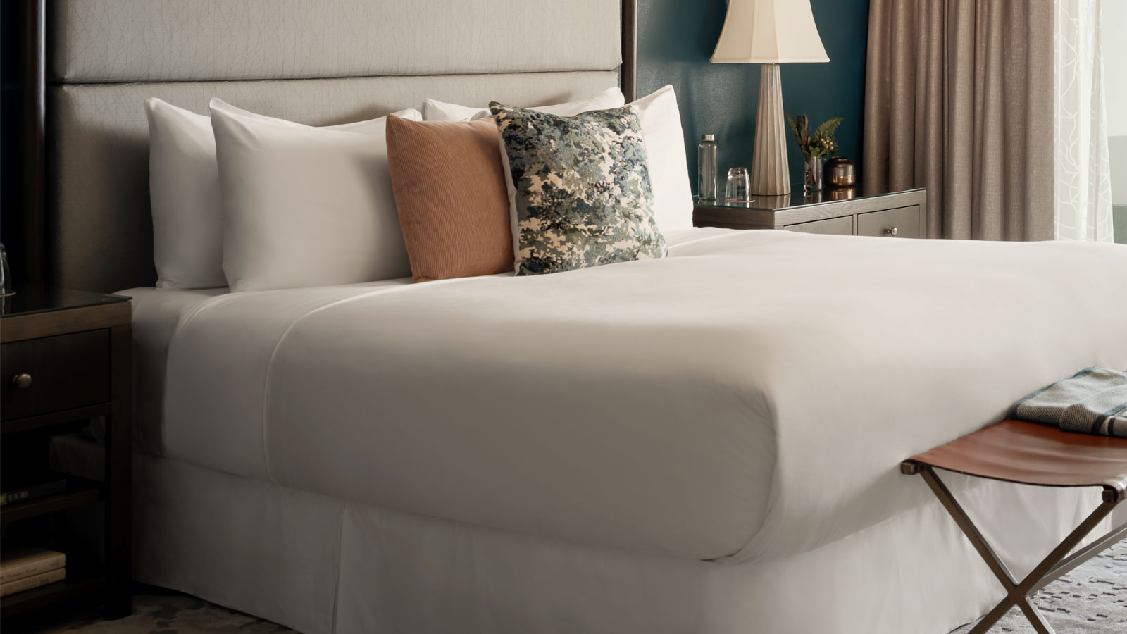 Close-up of a made bed with white linens, three decorative pillows, a bedside table with a lamp, and a small bench at the foot.