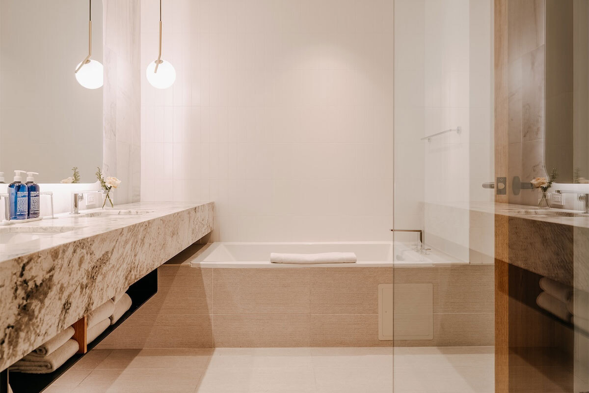 Modern bathroom with a beige tiled bathtub, marble countertop with sinks, white towel on tub edge, and hanging globe lights.