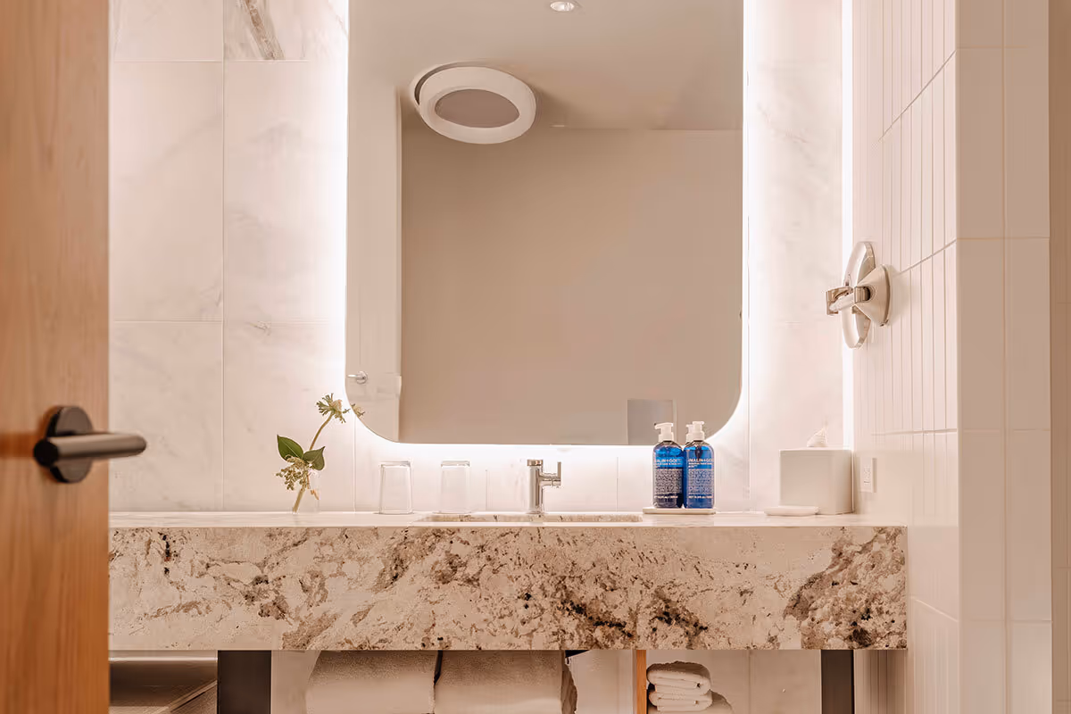 Modern bathroom with a large illuminated mirror, beige marble countertop, single sink, two blue soap dispensers, and neatly folded white towels underneath.