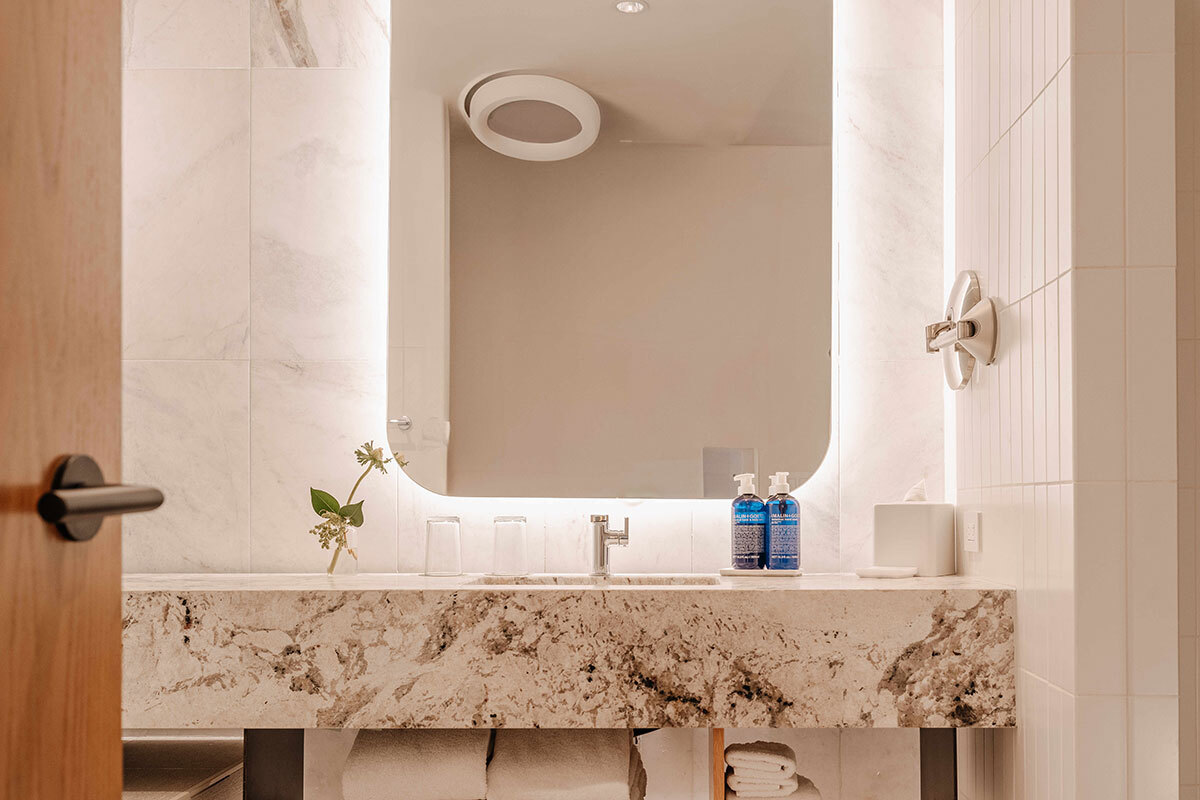 Modern bathroom with a large illuminated mirror, beige marble countertop, single sink, two blue soap dispensers, and neatly folded white towels underneath.