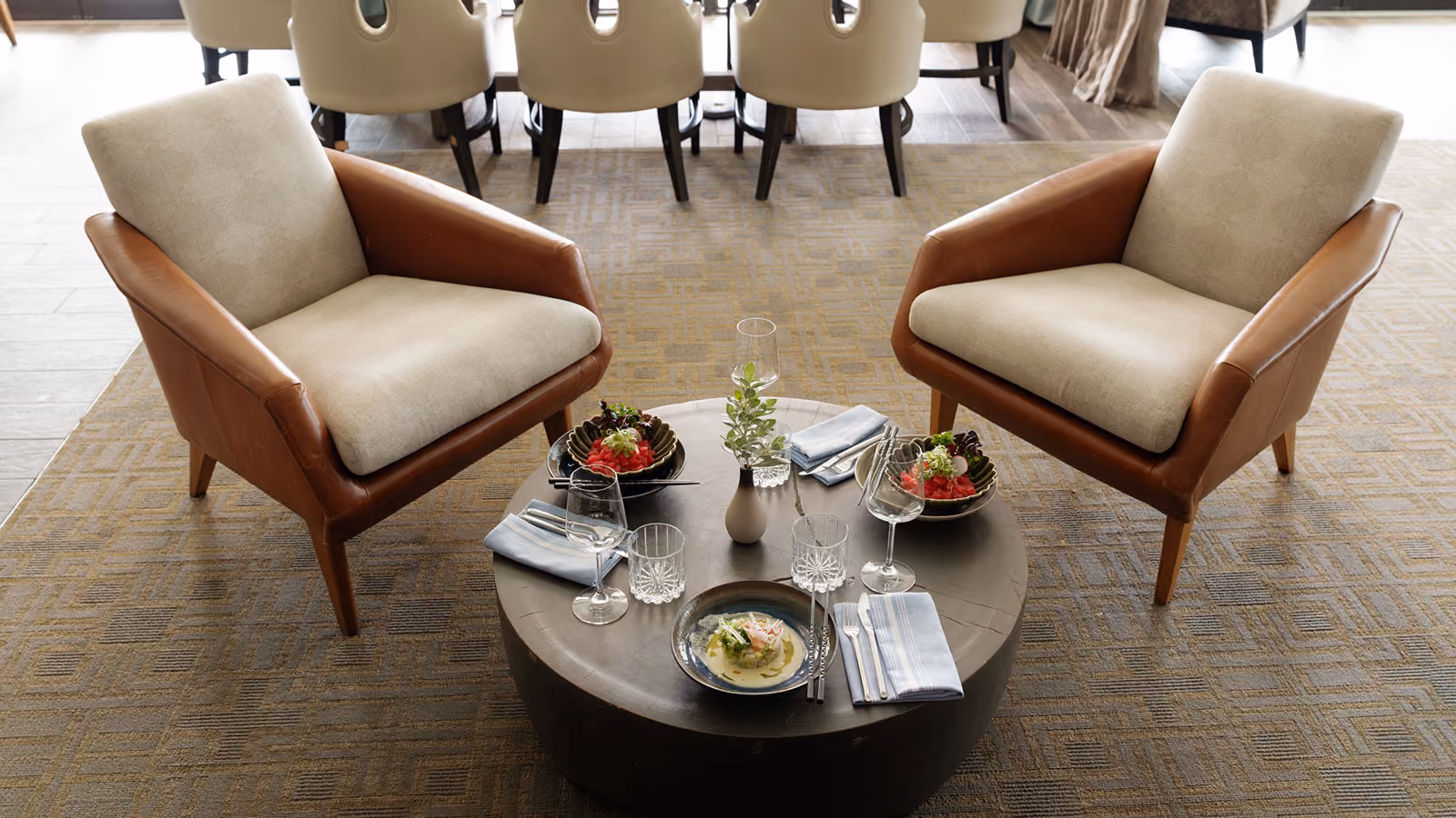 Two brown and beige armchairs facing a round table set with dishes, glasses, and napkins on a patterned rug.