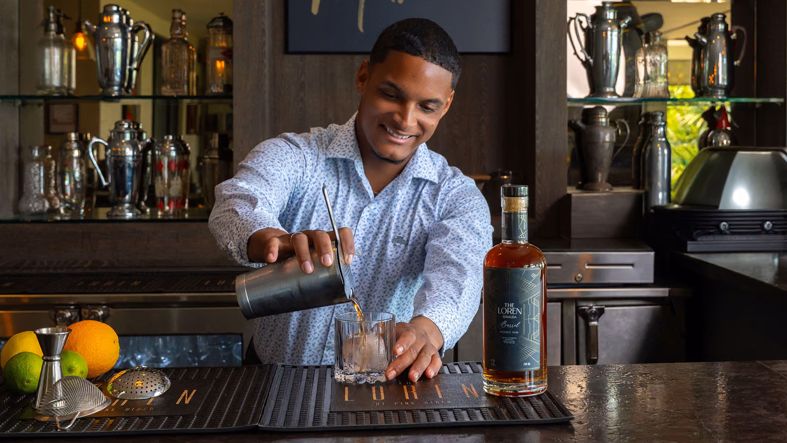 Smiling bartender pouring a drink from a shaker into a glass with a bottle of The Loren rum on the bar.