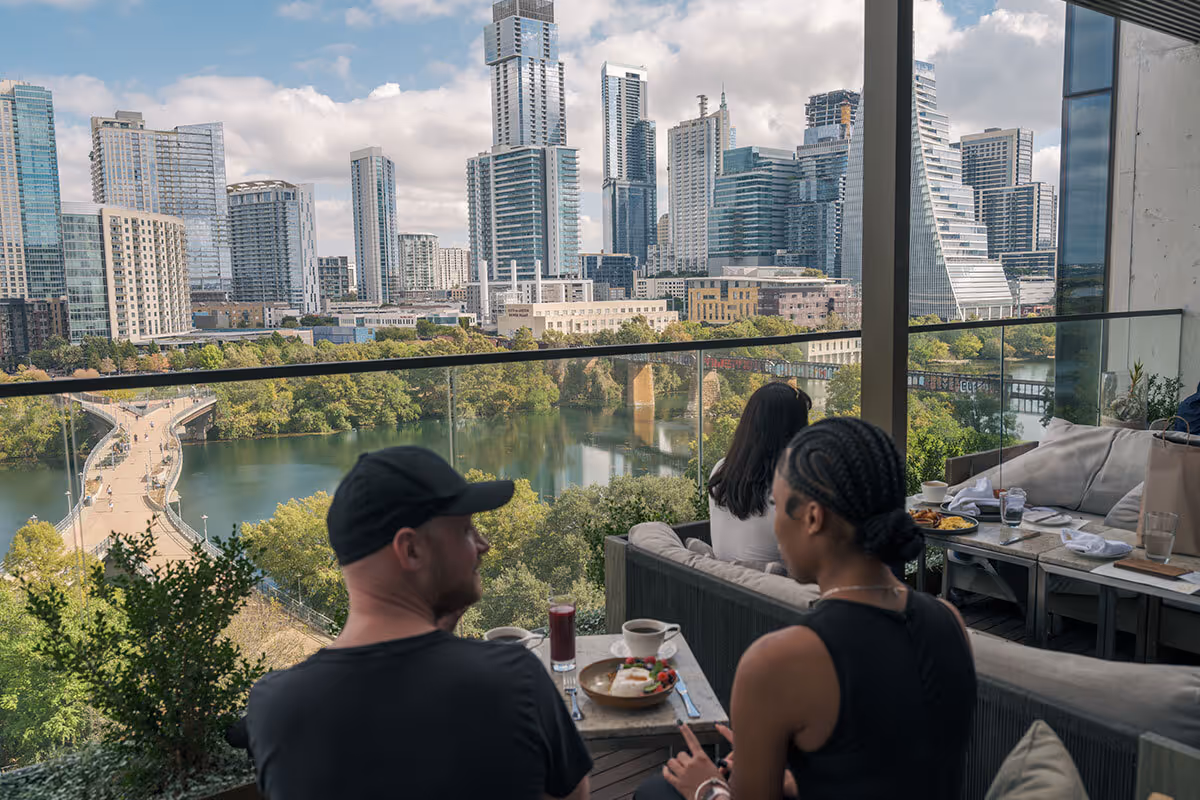 Two people dine on a terrace overlooking a river and the city skyline with tall modern buildings.