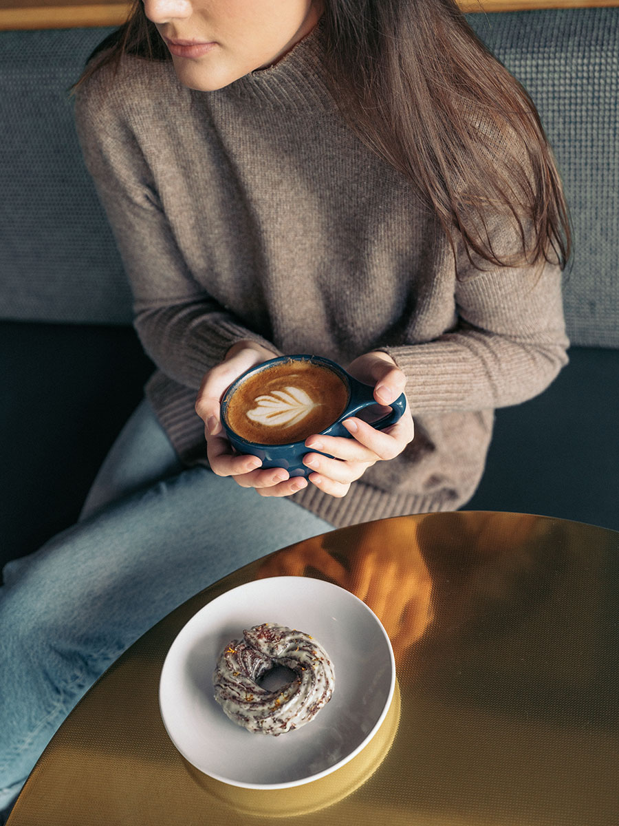 Person in a brown sweater holds a cup of latte with leaf art, with a pastry on a white plate on a gold table.