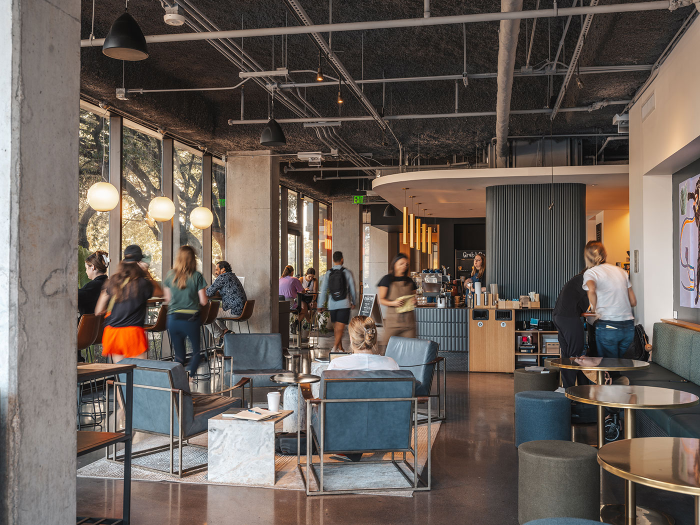 Modern coffee shop interior with people standing in line at the counter and others sitting in lounge chairs.