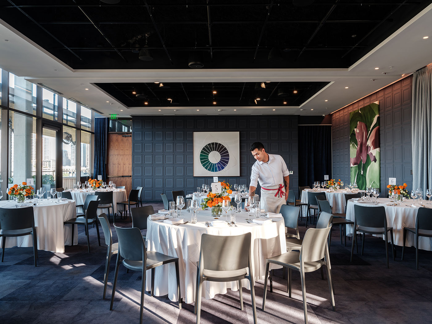 A waiter sets a round table in a modern, upscale dining room with neatly arranged tables and floral centerpieces.