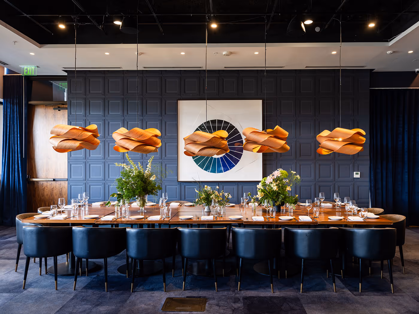 Long dining table with black chairs, set with plates and glasses, beneath modern pendant lights and geometric wall art.