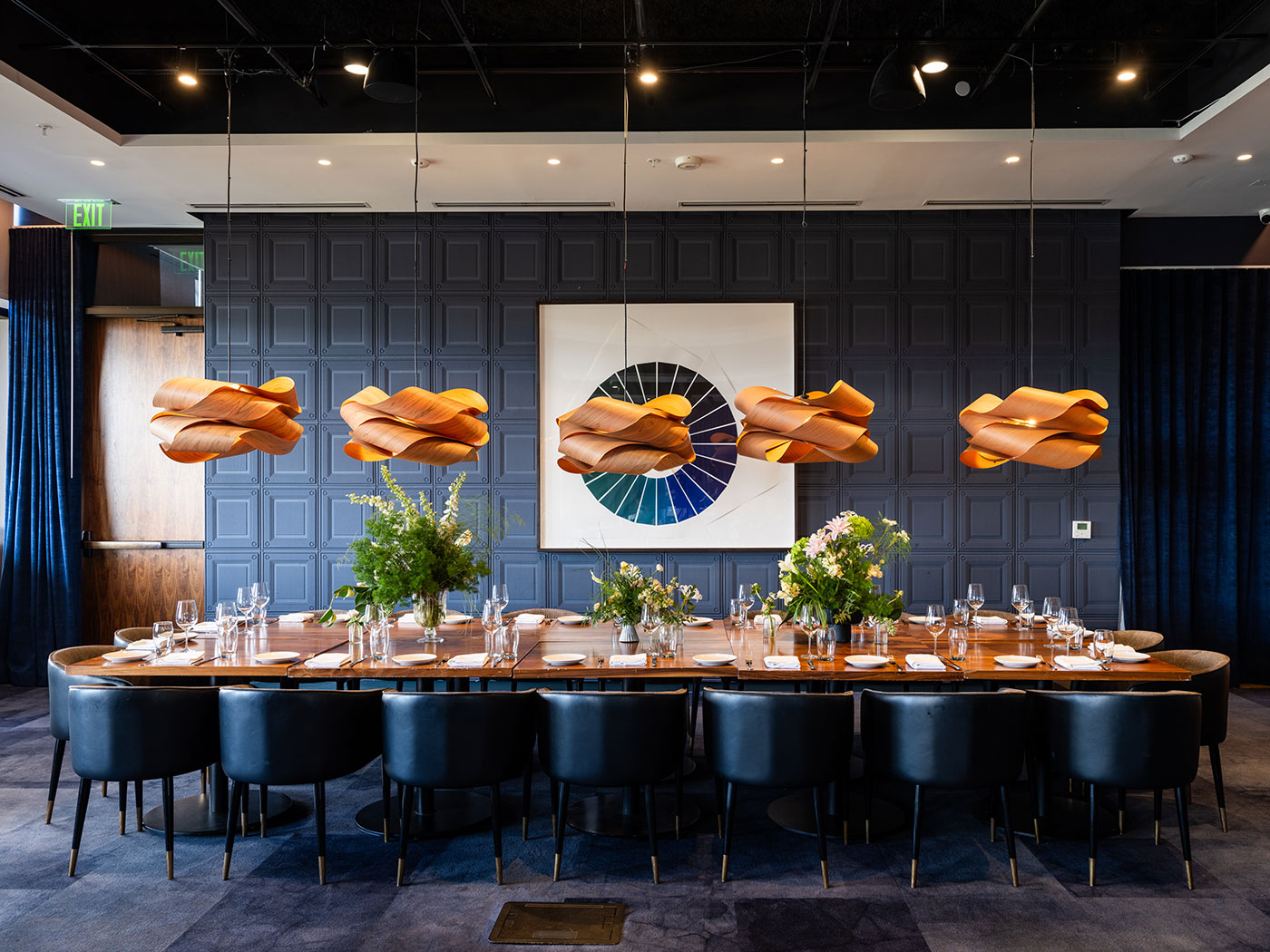 Long dining table with black chairs, set with plates and glasses, beneath modern pendant lights and geometric wall art.