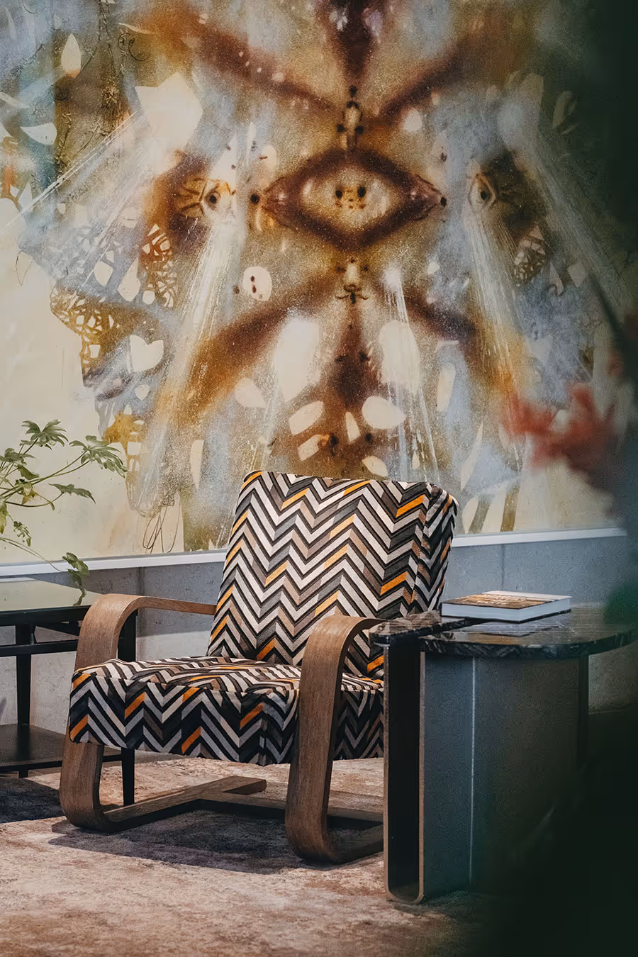 A patterned armchair sits by a side table in front of a mural featuring abstract eye and butterfly designs.