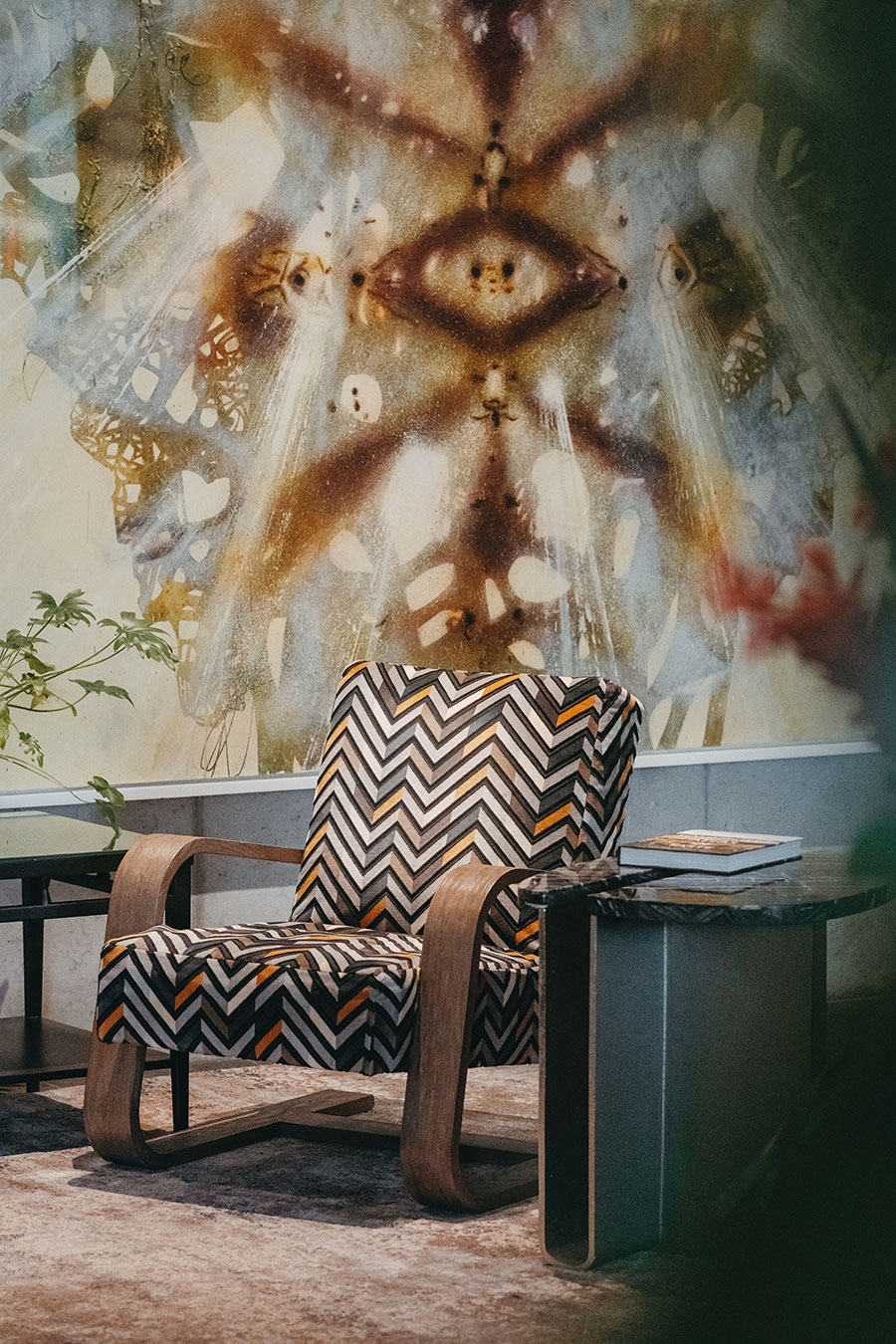 A patterned armchair sits by a side table in front of a mural featuring abstract eye and butterfly designs.