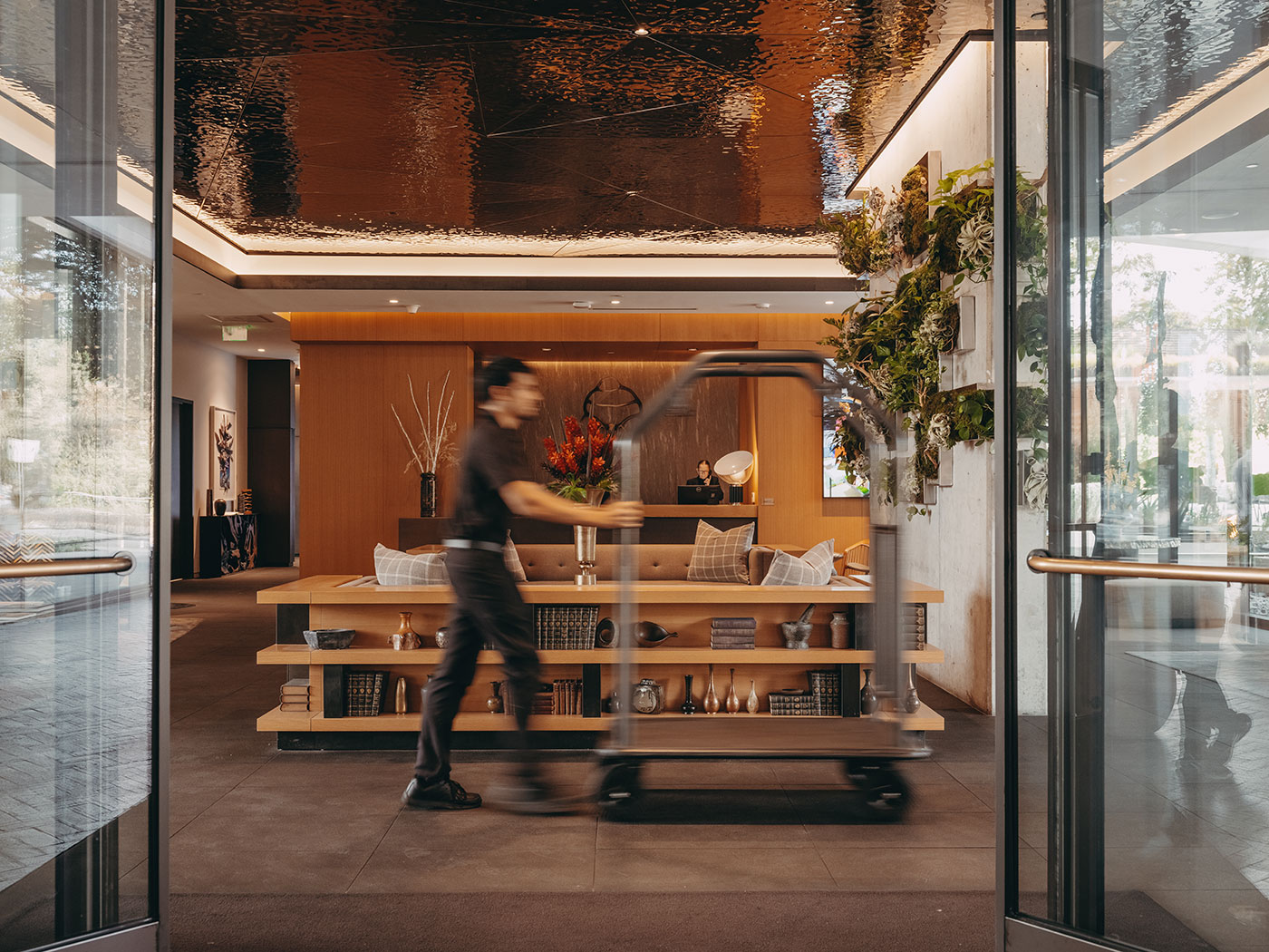 A person pushes a luggage cart through a modern hotel lobby with wooden decor and plants on the wall.