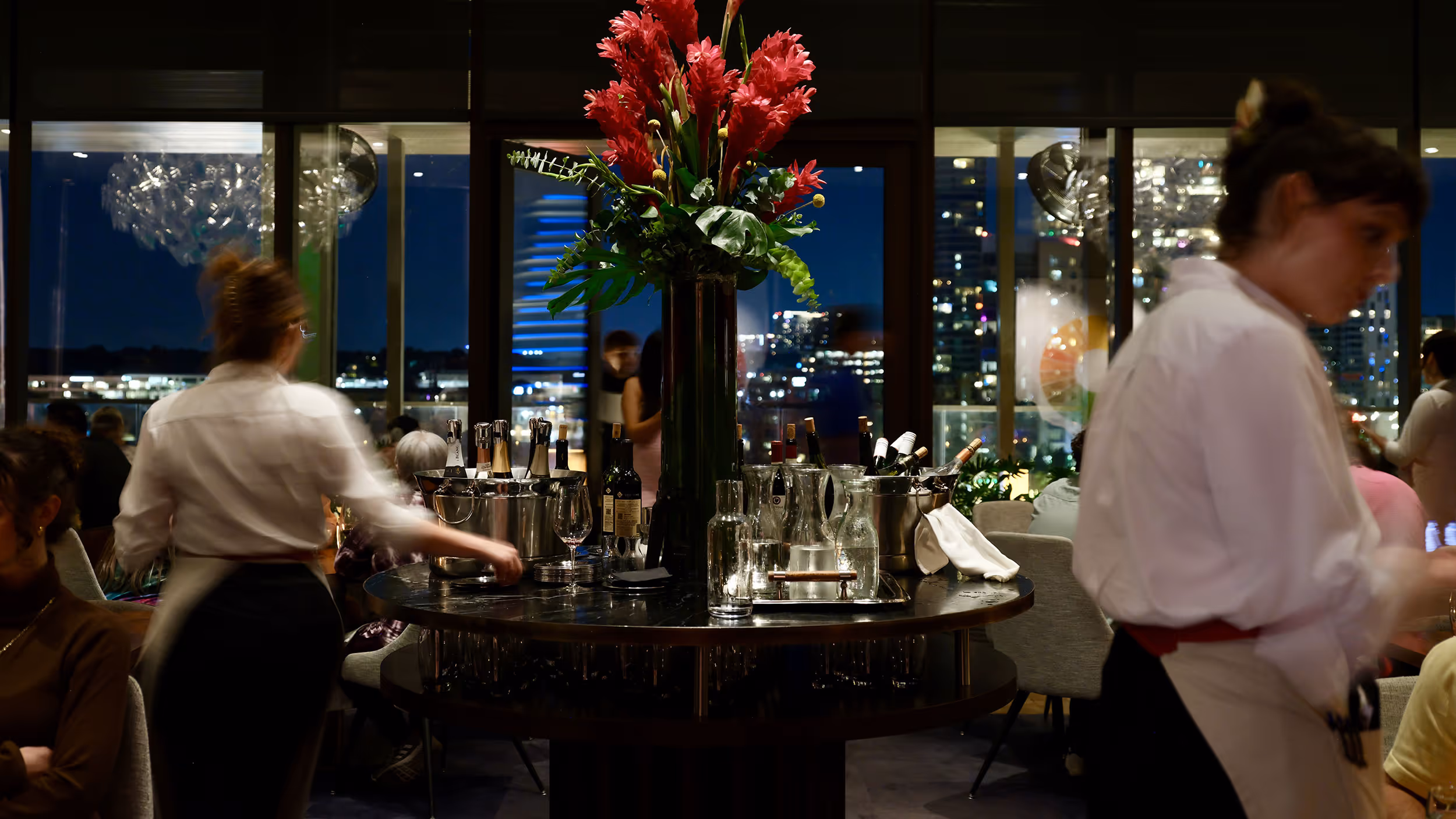 Waitstaff move around a round table with flower arrangement and bottles in a dimly lit restaurant with city views at night.