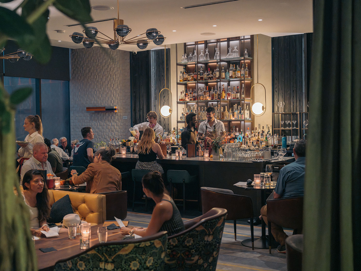 People dine and converse in a modern restaurant with a well-lit bar and bartenders preparing drinks in the background.