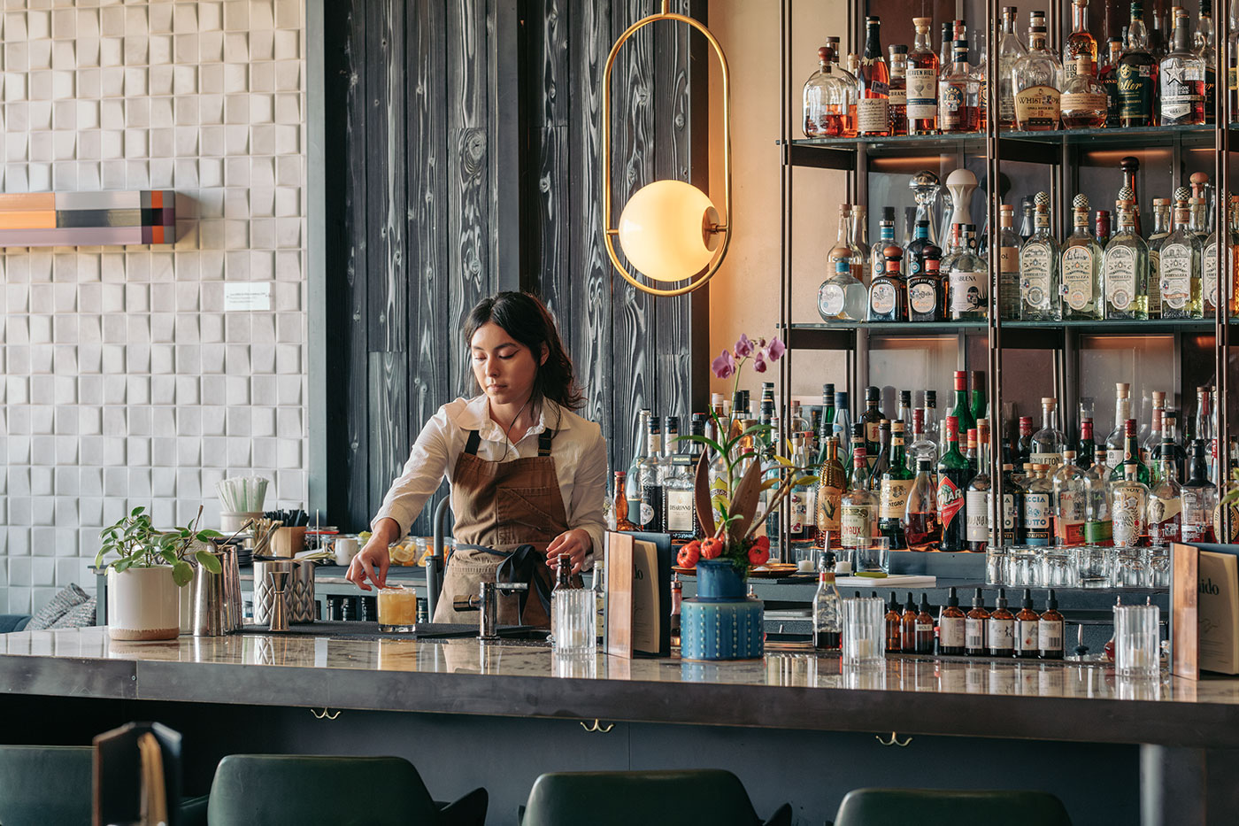 A bartender prepares a drink behind a modern bar stocked with bottles and glassware, with plants and decor on the counter.