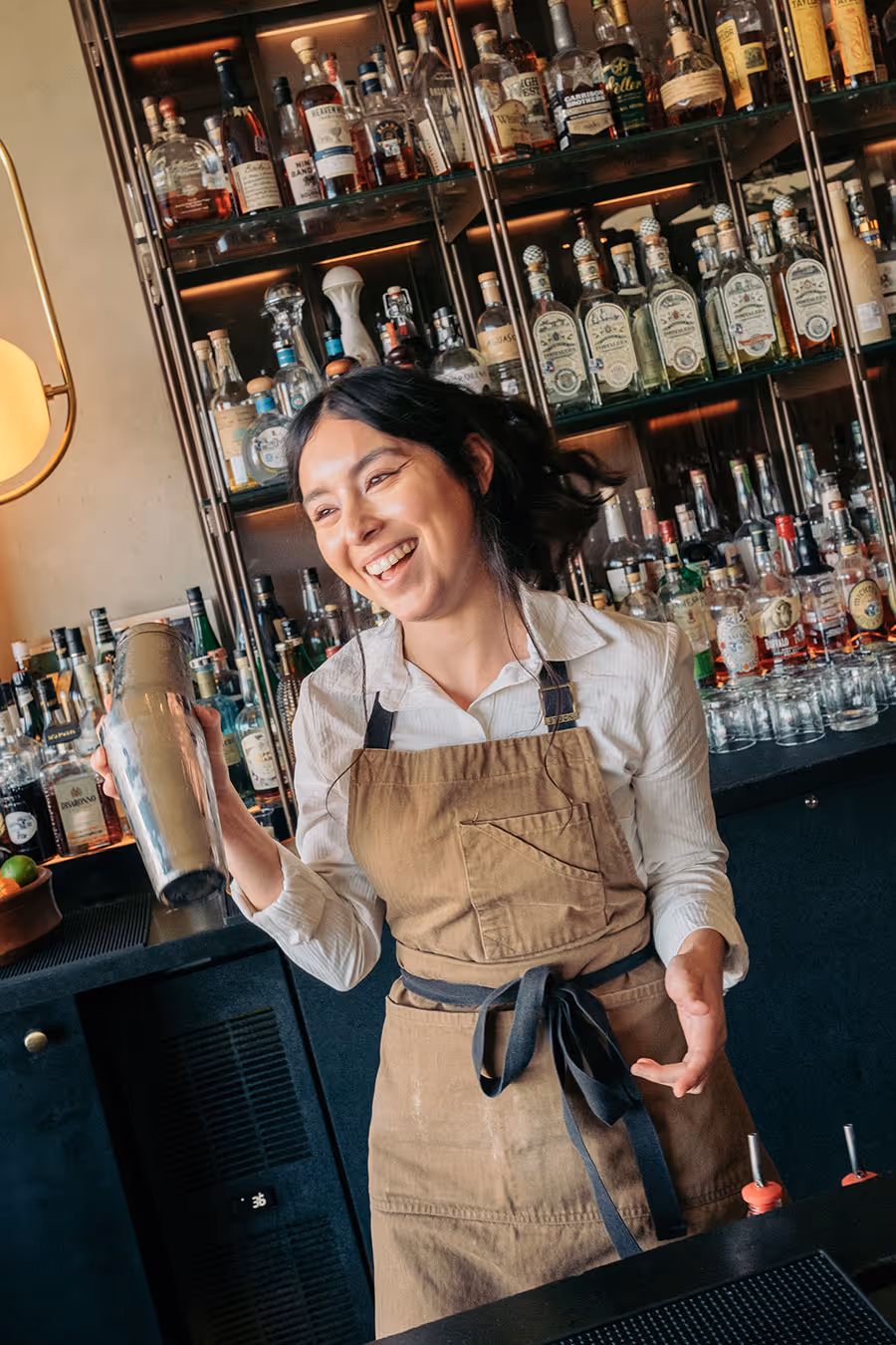 Bartender in apron smiling while shaking a cocktail shaker behind a bar stocked with bottles of liquor.