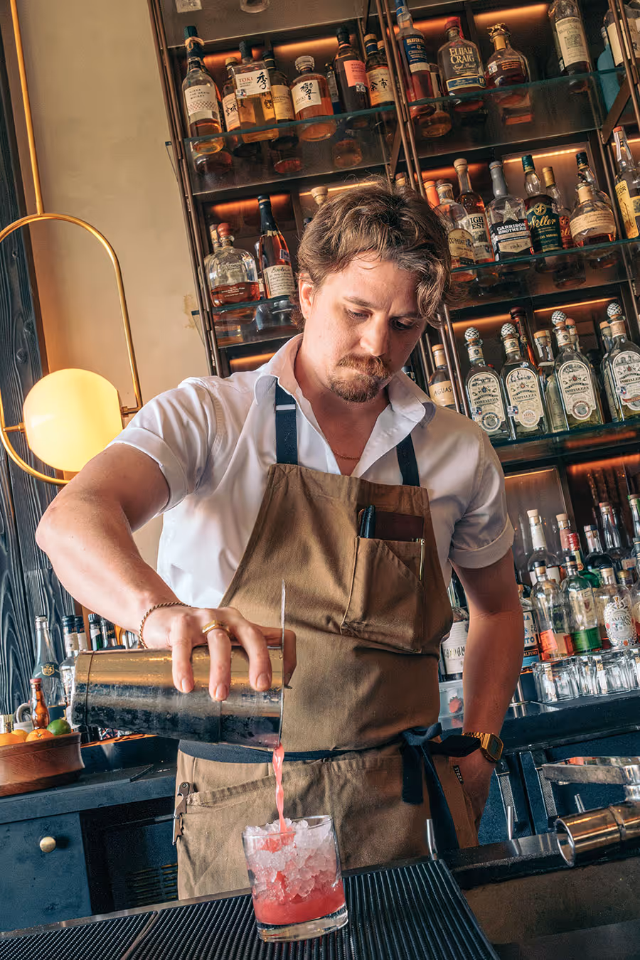 Bartender in a brown apron pours a red cocktail from a shaker into a glass filled with ice at a bar.