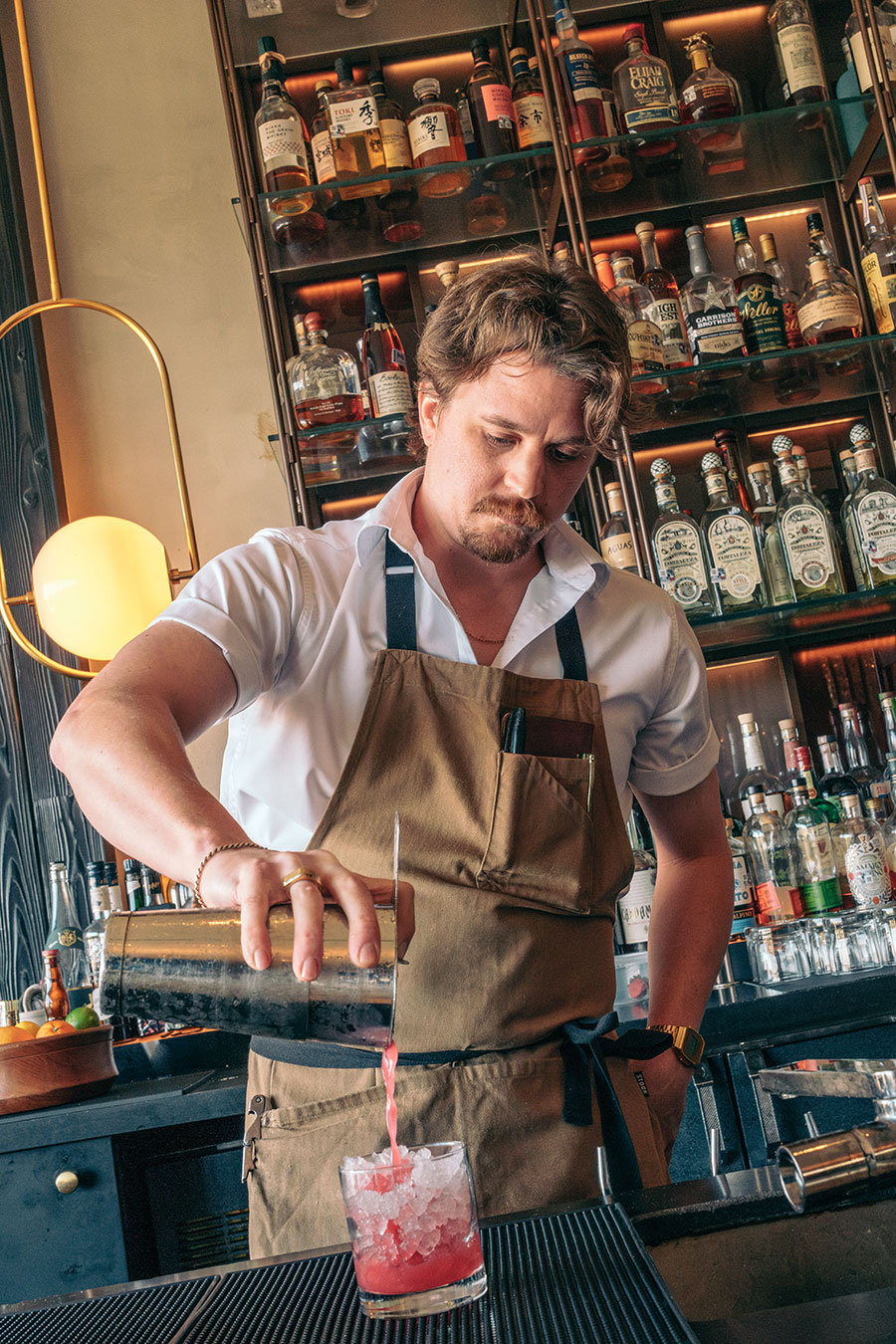 Bartender in a brown apron pours a red cocktail from a shaker into a glass filled with ice at a bar.