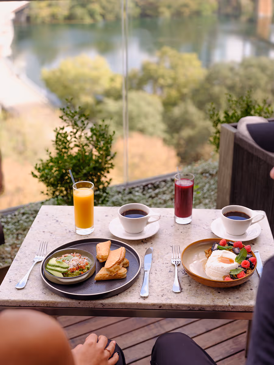 Two people sit at a table with coffee, juice, potato wedges, avocado toast, and a dessert topped with berries.