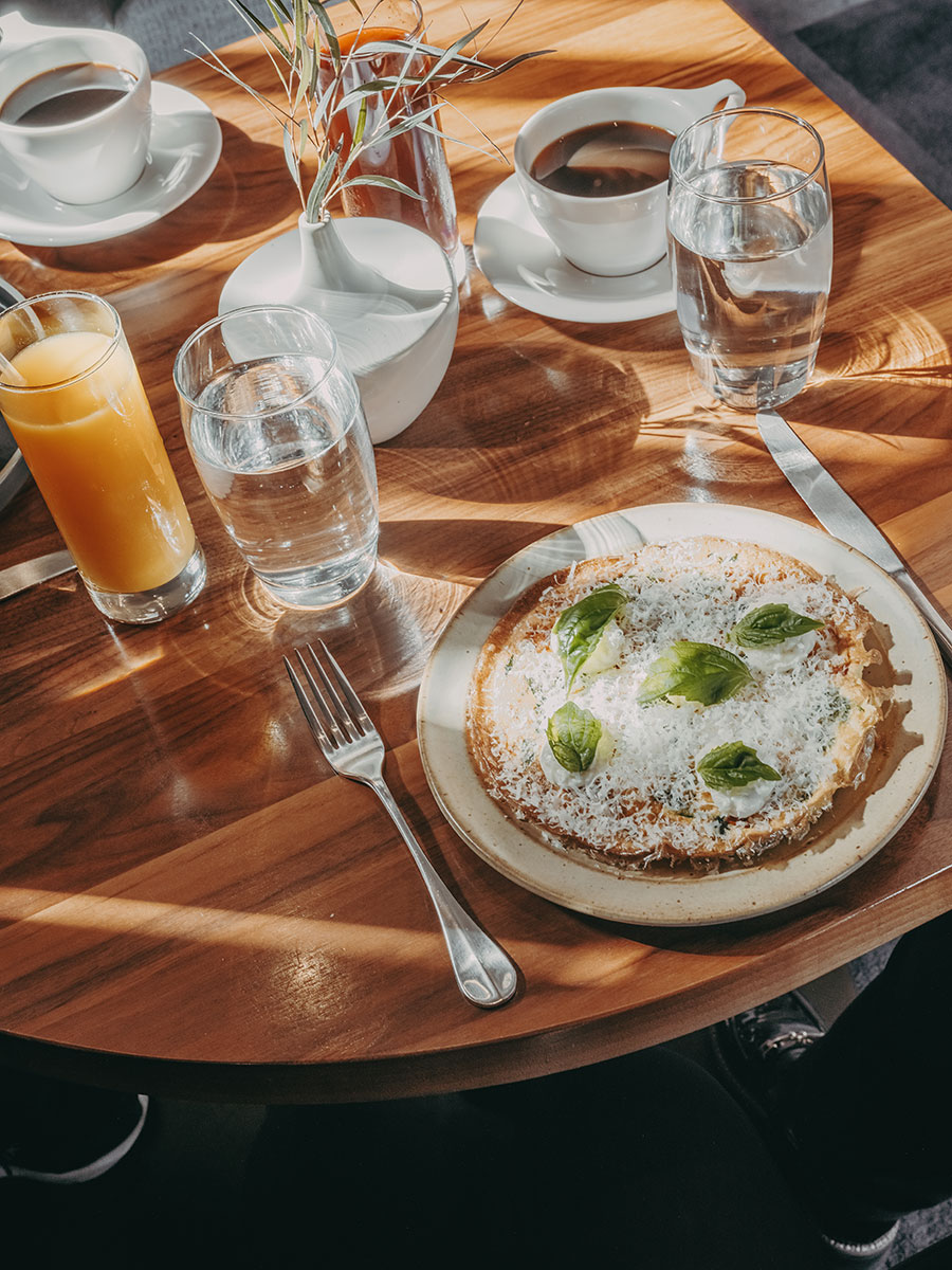 A plate with a pancake topped with cheese and basil, a fork and knife, water, orange juice, and coffee on a wooden table.