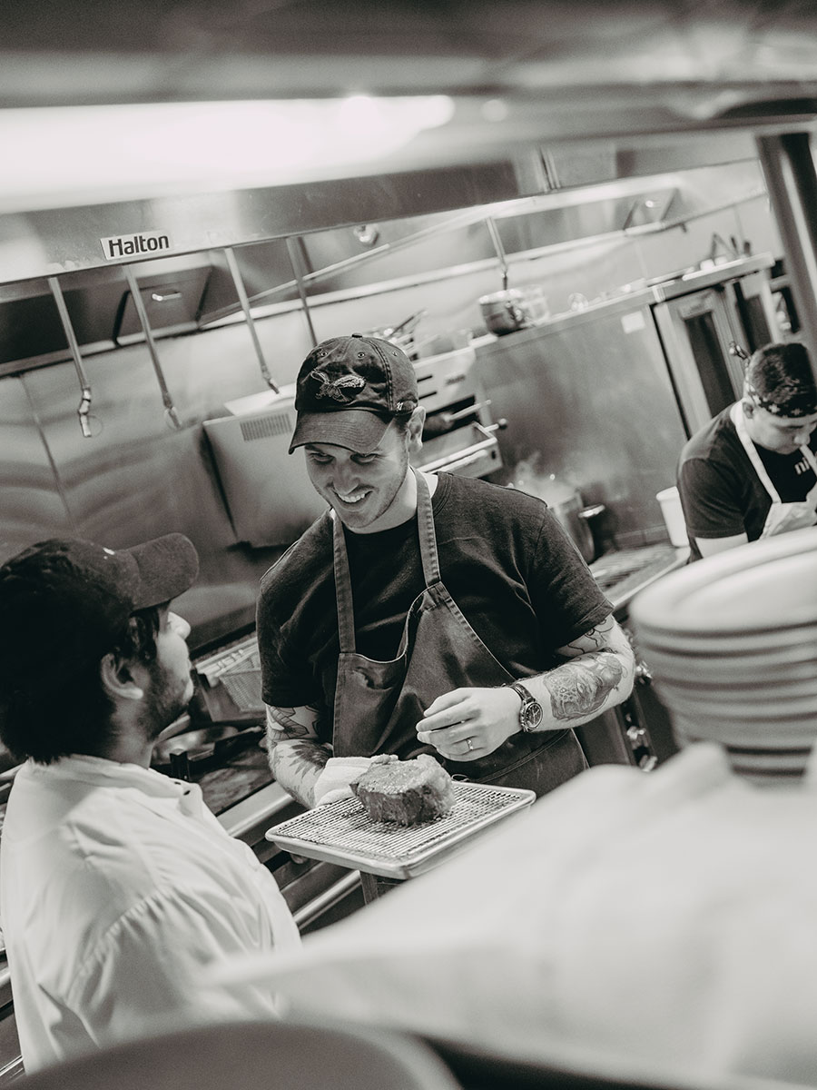 Two chefs talk and smile in a commercial kitchen; one holds a plated dish while a third chef works in the background.