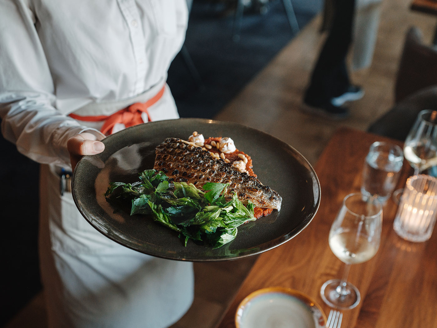 Server holding a plate with cooked fish, greens, and sauce next to a set table with wine glasses and a lit candle.