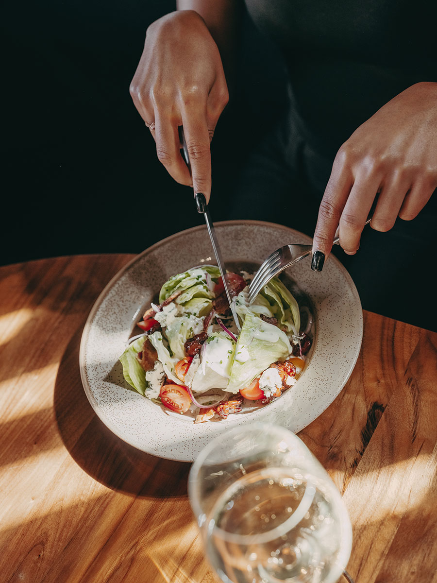 Person using fork and knife to eat a salad with lettuce, tomatoes, and cheese at a wooden table with a glass of water.