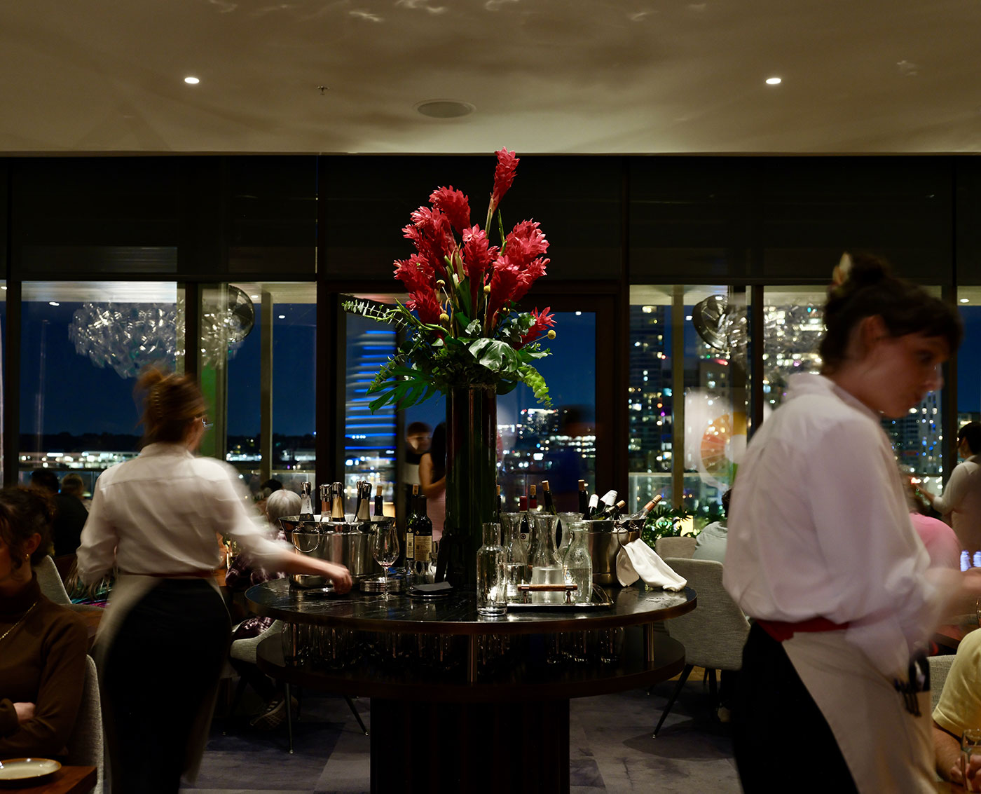 Restaurant interior at night with a large vase of red flowers on a central table, people dining and city lights visible outside.