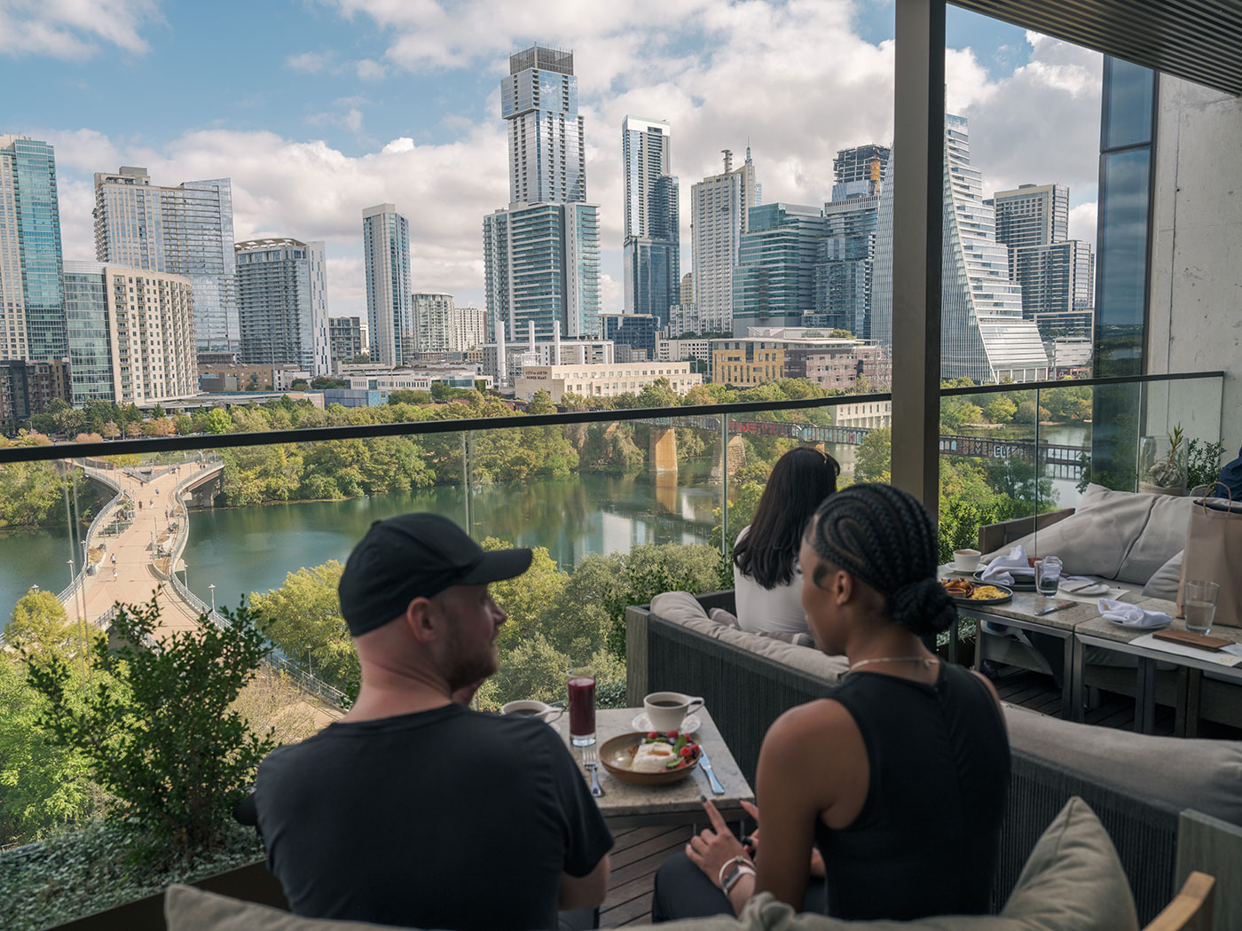 Two people dine on a terrace overlooking a river and a modern city skyline under a partly cloudy sky.