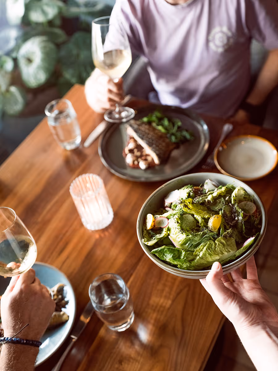 A person holds a bowl of salad at a wooden table set with wine, water glasses, and plates of food.