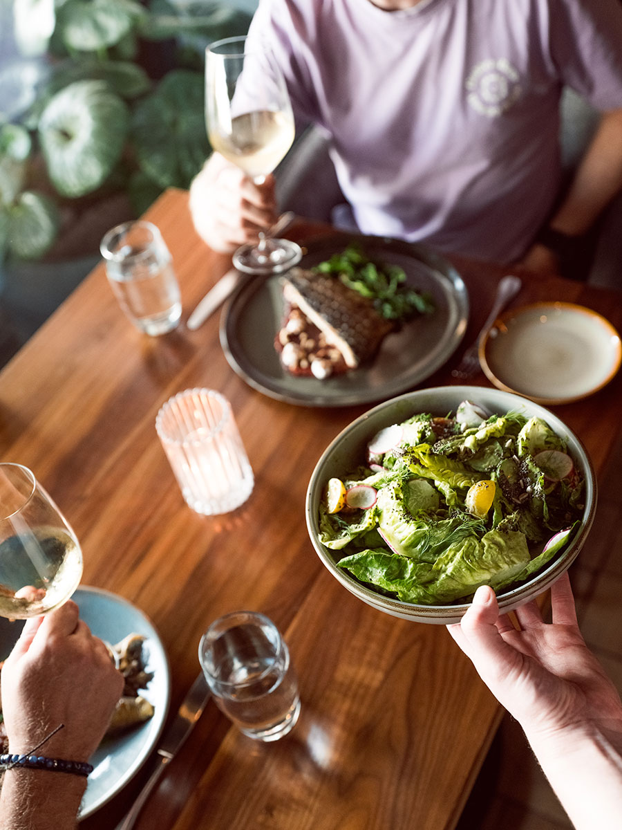 A person holds a bowl of salad at a wooden table set with wine, water glasses, and plates of food.
