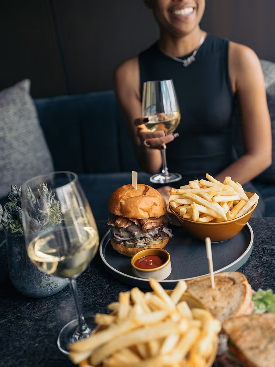 A person holding a glass of white wine sits at a table with a burger, fries, and another glass of wine.