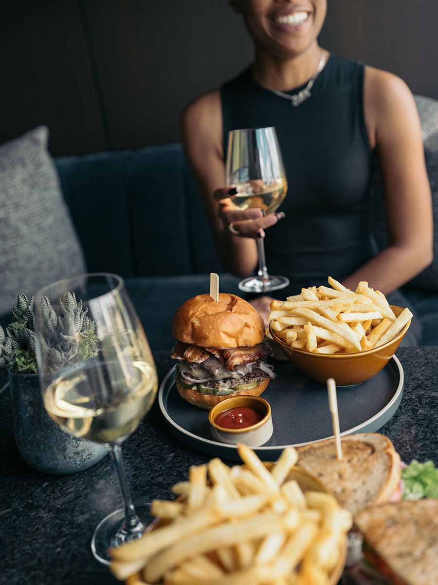 A person holding a glass of white wine sits at a table with a burger, fries, and another glass of wine.