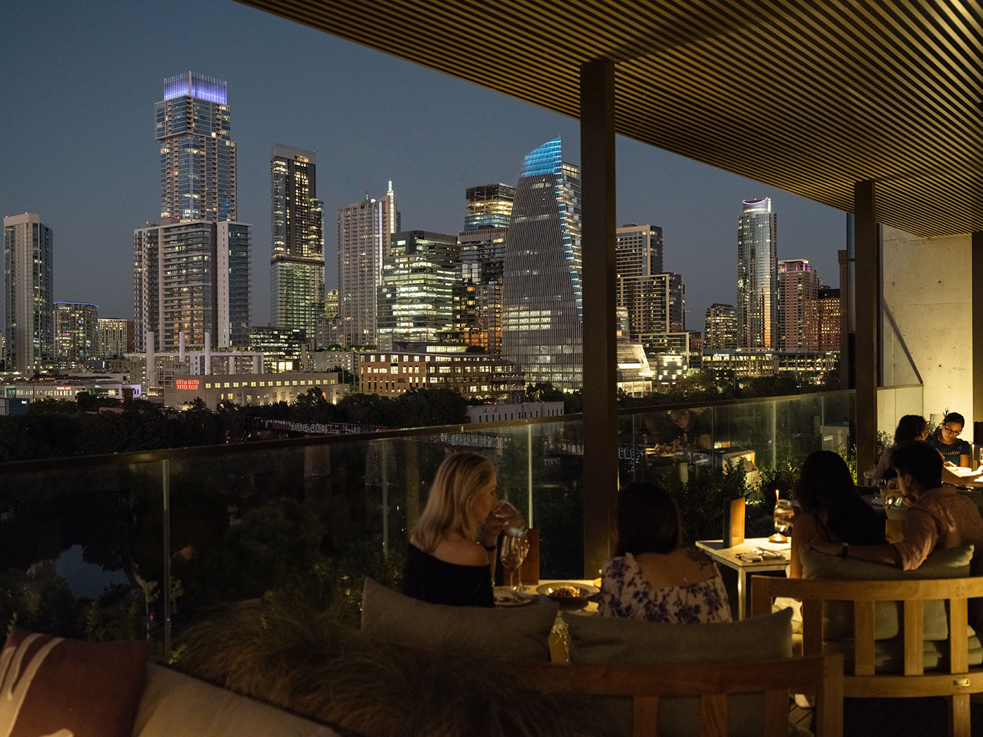 People dining on an outdoor terrace at night with a city skyline and tall buildings in the background.